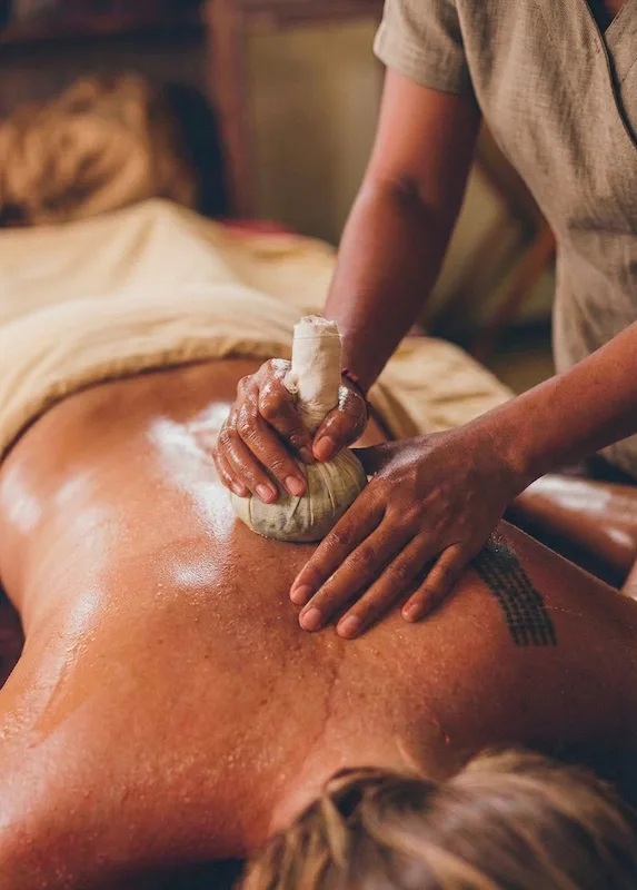 A professional spa therapist at Goddess Retreats Bali performs a traditional Elakkizhi massage using a heated medicinal herb poultice and warm botanical oils on a client's back for deep tissue restoration and joint relief.