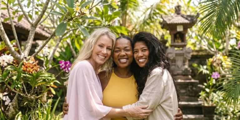 Three vibrant women in their 40s and 50s smiling together in a tropical Bali yemple garden while enjoying a holistic longevity women’s retreat.