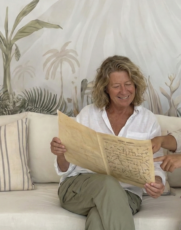 A smiling woman in her 60s sitting on a cozy rattan sofa at Goddess Retreats in Bali, reviewing a Vedic numerology and astrology chart during a soul-alignment session. A practitioner's hands point to the document as she sits in front of a tropical volcano mural, experiencing clarity and personal growth at a women's healing retreat.